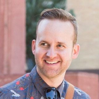 A man in a blue shirt with red hearts and a brown strap smiles warmly against a vibrant red backdrop.